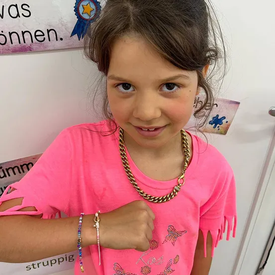 Ein junges Mädchen mit braunen Haaren, das ein rosa T-Shirt und eine goldene Halskette trägt, posiert für ein Foto mit einer weißen Wand im Hintergrund.