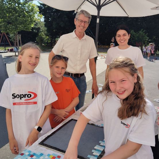 Drei Kinder und zwei Erwachsene stehen vor einem Tisch mit einer Mosaikplatte. Sie lächeln alle und scheinen für ein Foto zu posieren. Sie befinden sich in einem Außenbereich.