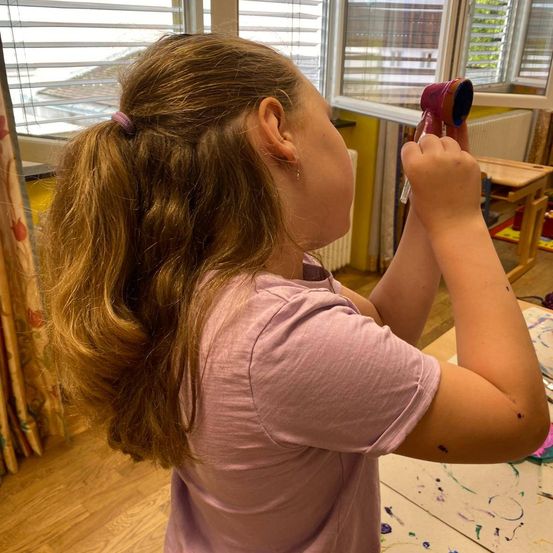 Ein junges Mädchen mit langen blonden Haaren hält ein buntes Spielzeug und bläst hinein. Sie steht vor einem Tisch mit Farbspritzern und schaut aus dem Fenster.