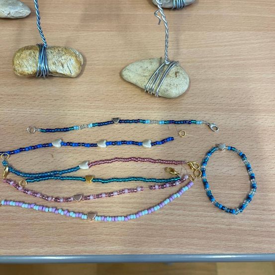Verschiedene Perlenketten und Armbänder mit Herzanhängern sind auf einer Holzoberfläche ausgestellt. Zwei mit Draht umwickelte Steine befinden sich hinter dem Schmuck.