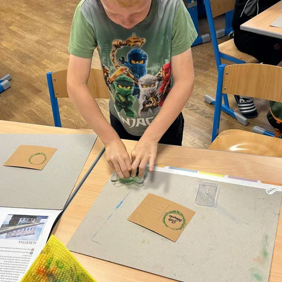 Ein Junge arbeitet in einem Klassenzimmer an einem Bastelprojekt am Tisch. Er benutzt einen Stempel auf einem Stück Pappe. Andere Materialien und Stühle sind um ihn herum.