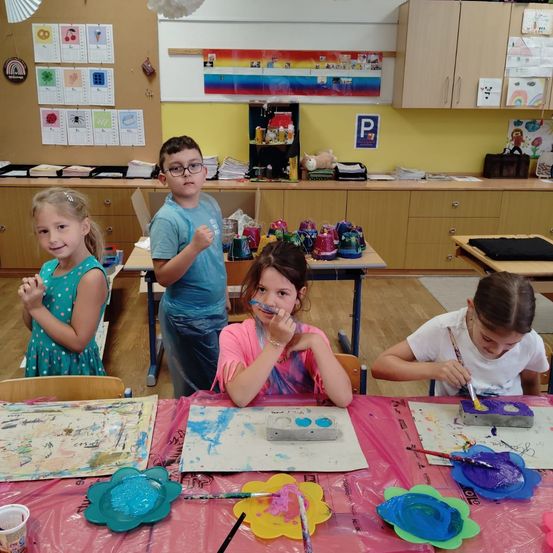 Vier Kinder malen an einem Tisch im Klassenzimmer. Eines benutzt einen Malkasten und Pinsel. Ein anderes steht und trägt eine Brille. Ein drittes sitzt und malt auf eine Leinwand. Das letzte Kind sitzt und malt auf einen Tonblock.
