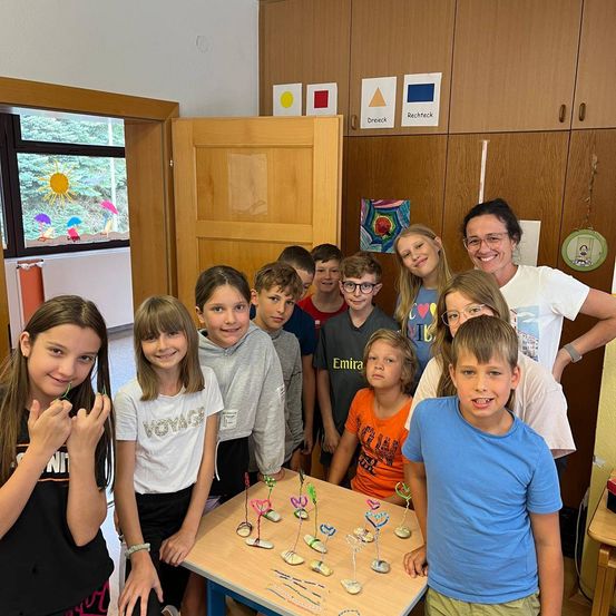 Eine Gruppe von Kindern und einer erwachsenen Frau stehen in einem Klassenzimmer, lächeln und posieren für ein Foto. Ein Tisch mit herzförmigen Objekten steht vor ihnen. Dahinter befinden sich Holzschränke mit bunten geometrischen Formen.