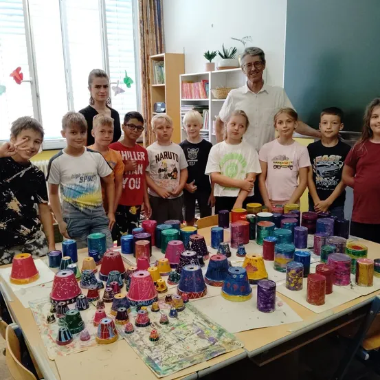 Eine Gruppe von Kindern und Erwachsenen steht in einem Klassenzimmer und lächelt für ein Foto. Sie umgeben einen Tisch, der mit bunt bemalten Bechern bedeckt ist. Ein Mann steht hinten bei einer Frau.