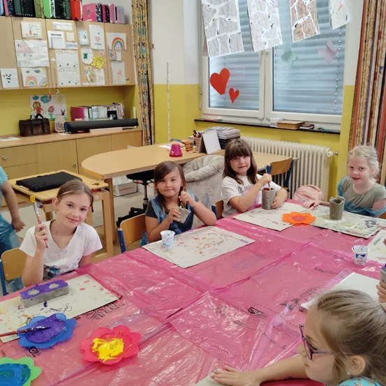 Fünf junge Mädchen sitzen in einem Klassenzimmer an einem Tisch und malen. Sie haben Farbe an den Händen und im Gesicht. Der Tisch ist mit einer rosa Tischdecke bedeckt. Dahinter befinden sich Schränke und ein Heizkörper. Das Fenster hat Zeichnungen und Herzen.