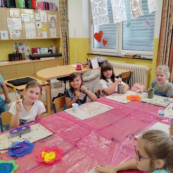 Fünf junge Mädchen sitzen in einem Klassenzimmer an einem Tisch und malen. Sie haben Farbe an den Händen und im Gesicht. Der Tisch ist mit einer rosa Tischdecke bedeckt. Dahinter befinden sich Schränke und ein Heizkörper. Das Fenster hat Zeichnungen und Herzen.