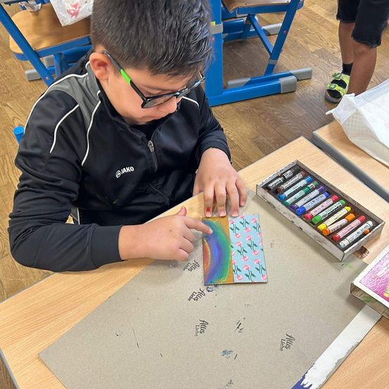 Ein Junge sitzt an einem Schreibtisch und malt ein kleines Stück Papier mit Wasserfarben. Eine Box mit Buntstiften und ein weiteres Blatt Papier liegen auf dem Schreibtisch.