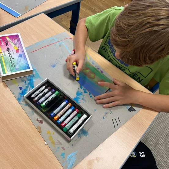 Ein Junge in einem grünen T-Shirt malt auf einem Schreibtisch mit Aquarellstiften und einer Palette.