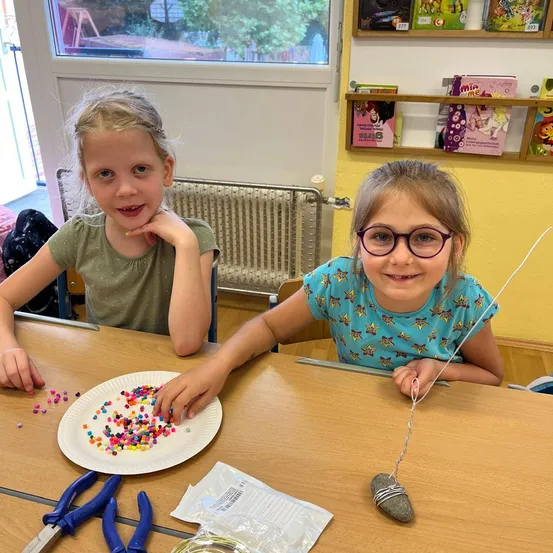 Zwei Mädchen in einem Klassenzimmer mit einem Tisch, sie basteln. Eines hält eine Schnur und einen Stein, das andere spielt mit bunten Perlen auf einem Teller. Werkzeuge und Bücher sind in der Nähe.