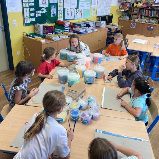 Ein Klassenzimmer mit Schülern an Tischen, die eine Bastelaktivität mit verschiedenen Materialien durchführen. Bunte Behälter halten verschiedene farbige Substanzen. Der Raum ist gut beleuchtet mit Regalen voller Bücher.