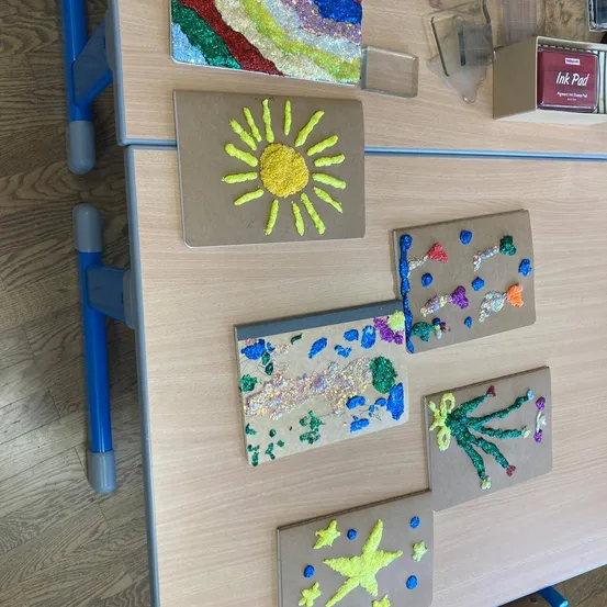 Ein Holztisch zeigt verschiedene farbenfrohe Kunstwerke, darunter eine Sonne mit gelber Wolle, ein abstraktes farbenfrohes Stück, eine fischartige Kreation und eine handartige Kunst mit grüner Wolle.