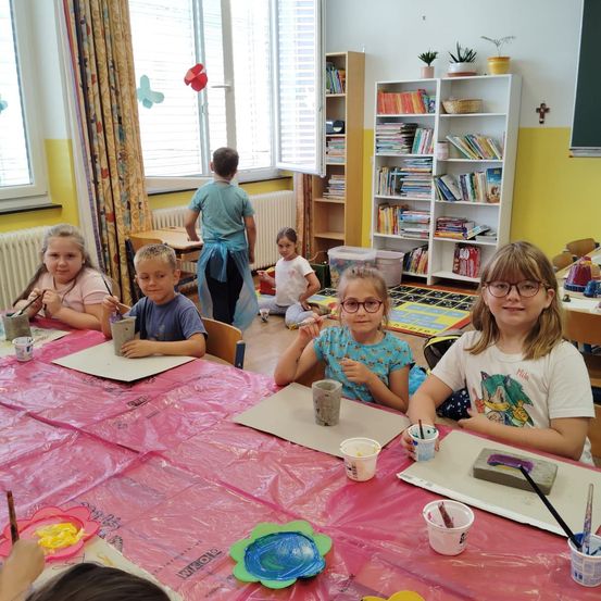 Mehrere Kinder sitzen in einem Klassenzimmer um einen Tisch herum und tragen eine Brille und eine Schürze. Sie sind in einer Töpferaktivität engagiert. Der Tisch ist mit einer Plastikfolie abgedeckt. Verschiedene Kunstutensilien, einschließlich Pinsel, Farbbecher und Ton, sind auf dem Tisch platziert. Im Hintergrund befinden sich mit Büchern gefüllte Bücherregale.