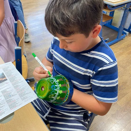Ein junger Junge sitzt in einem Klassenzimmer an einem Schreibtisch und zeichnet mit einem grünen Marker auf ein zylindrisches Objekt. Er trägt ein blau-weiß gestreiftes T-Shirt.