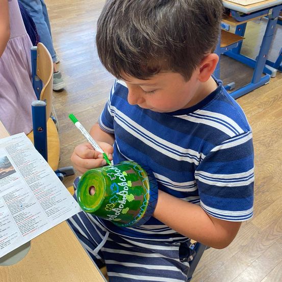 Ein junger Junge sitzt in einem Klassenzimmer an einem Schreibtisch und zeichnet mit einem grünen Marker auf ein zylindrisches Objekt. Er trägt ein blau-weiß gestreiftes T-Shirt.