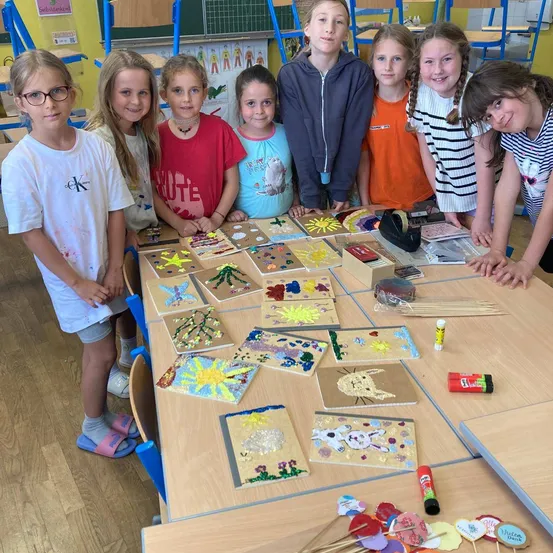 Eine Gruppe von Kindern steht um einen Tisch mit verschiedenen Kunstprojekten. Sie lächeln und schauen zur Kamera. Der Tisch ist mit bunten Zeichnungen, Papier und anderen Materialien gefüllt. Der Raum hat Holzböden und gelbe Wände.