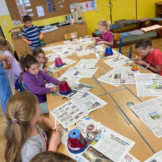 Eine Gruppe von Kindern sitzt an einem Tisch in einem Klassenzimmer und ist mit einer Bastelaktivität beschäftigt. Sie dekorieren Becher mit Farbe und Zeitungsausschnitten. Einige Kinder haben ihre Kunstwerke fertiggestellt, während andere noch arbeiten. Ein Junge steht hinter dem Tisch und hält einen fertigen Becher. Das Klassenzimmer hat Holztische, eine gelbe Wand und verschiedene Gegenstände in den Regalen.