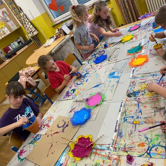Eine Gruppe von Kindern sitzt um einen Tisch und malt. Jedes Kind hat einen Pinsel und erschafft bunte Kunstwerke auf Papier und blumenförmigen Tellern.