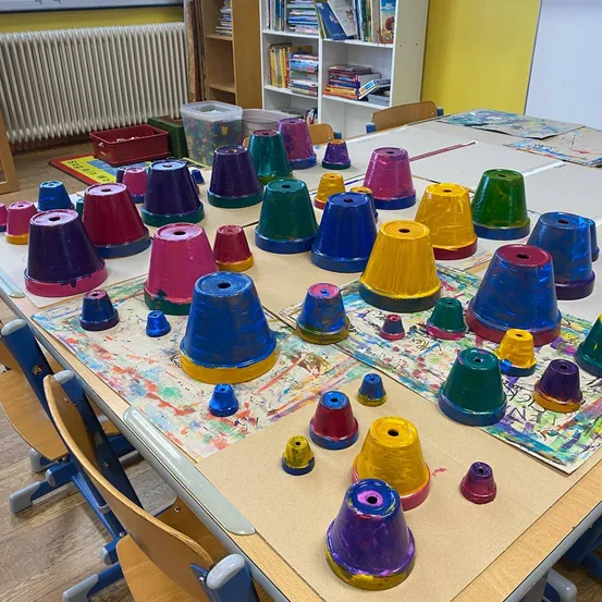 Ein Klassenzimmertisch, der mit verschiedenen farbigen Bechern auf Papier bedeckt ist. Stühle und Regale mit Büchern im Hintergrund.