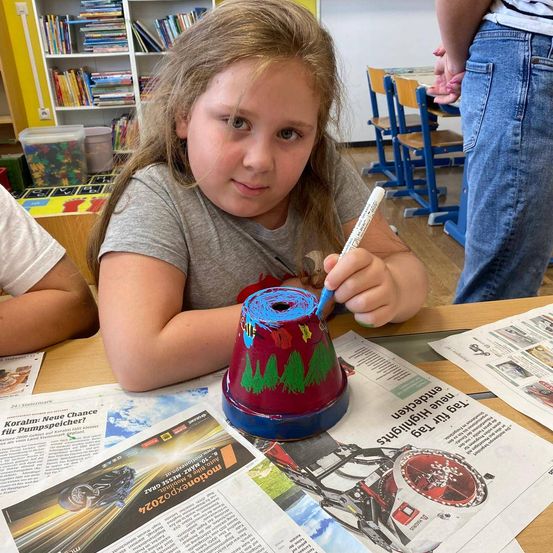 Ein kleines Mädchen in einem Klassenzimmer malt einen roten Topf mit blauer Farbe. Sie sitzt an einem Schreibtisch mit Zeitungen und Zeitschriften um sie herum.