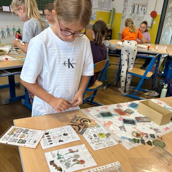 Ein Klassenzimmer mit Kindern, die an einem Kunstprojekt arbeiten. Ein Mädchen in einem weißen T-Shirt konzentriert sich auf ihr Projekt, während andere dahinter an ihren Projekten arbeiten.