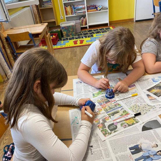 Drei Mädchen sind an einem Schreibtisch mit einer Aktivität beschäftigt und färben eine Glocke mit Markern. Der Schreibtisch ist mit Zeitungen und Markern übersät. Im Hintergrund ist eine Klassenzimmerumgebung zu sehen, mit Regalen, einem Teppich und einem Heizkörper.