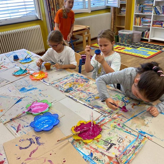 Vier junge Mädchen malen in einem Klassenzimmer. Drei von ihnen sitzen um einen Tisch herum, jede mit Pinsel und Bechern. Die vierte steht hinter ihnen und beobachtet. Der Tisch ist mit Papier und Farbspritzern bedeckt. Das Klassenzimmer hat gelbe Wände, Heizkörper, einen Teppich und Regale mit Büchern.