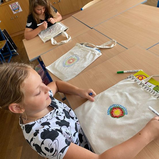 Zwei junge Mädchen in einem Klassenzimmer, die auf weiße Taschen zeichnen. Ein Mädchen hat die Augen geschlossen, während das andere auf einer Tasche mit Sonnenblumendesign zeichnet.