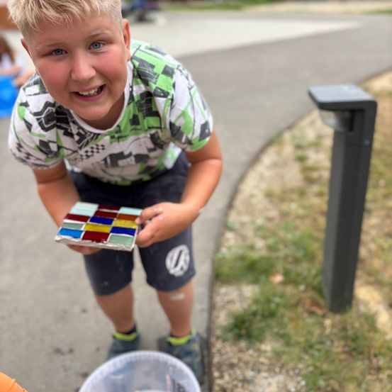 Ein junger Junge mit blauen Augen lächelt, während er ein farbenfrohes Mosaik-Fliese in den Händen hält und auf einem Bürgersteig steht.