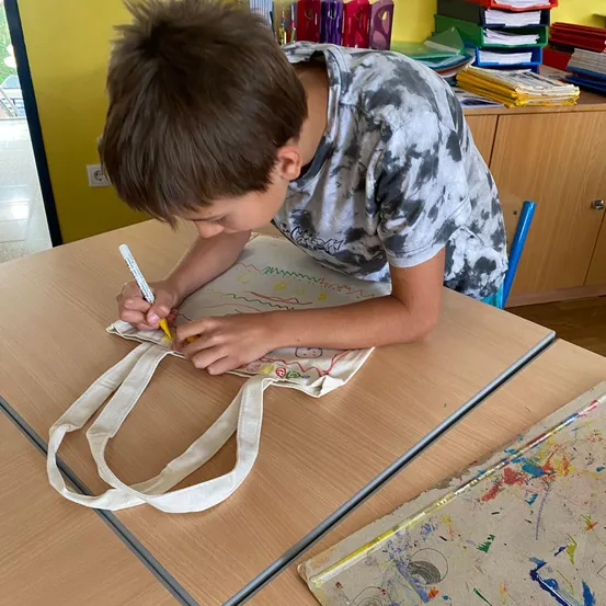 Ein Junge in einem Tie-Dye-Shirt zeichnet mit einem gelben Marker auf eine weiße Tragetasche. Er sitzt an einem Holzschreibtisch.
