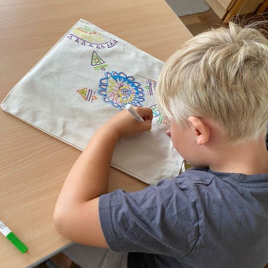 Ein Junge sitzt an einem Holztisch und malt ein weißes Tuch mit einem Marker. Das Tuch hat verschiedene geometrische Muster und Designs.