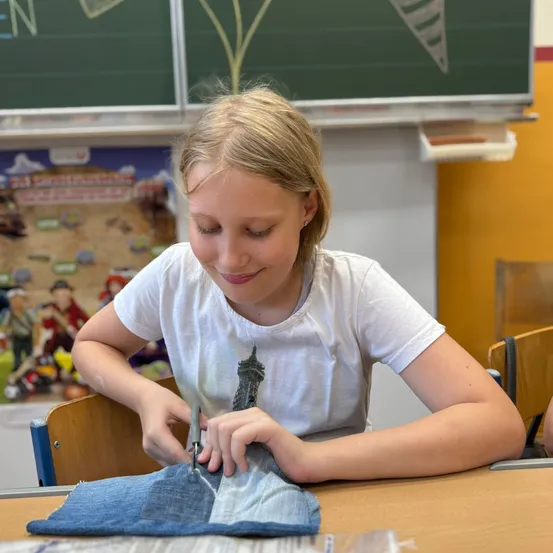 Ein junges Mädchen mit blonden Haaren sitzt an einem Schreibtisch und lächelt, während es ein Stück Jeansstoff mit einer Schere schneidet. Hinter ihr ist eine Klassenzimmerumgebung mit einer Tafel und einem bunten Poster zu sehen.