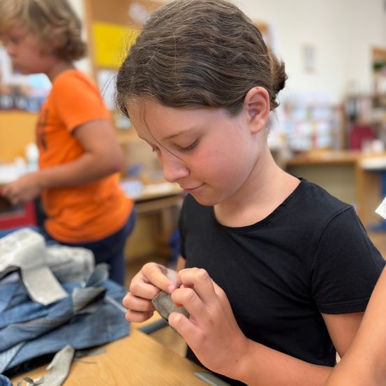 Ein junges Mädchen in einem schwarzen T-Shirt arbeitet an einem Steinstück, während ein anderes Kind in einem orangefarbenen T-Shirt hinter ihr steht. Sie sind in einem Klassenzimmer.
