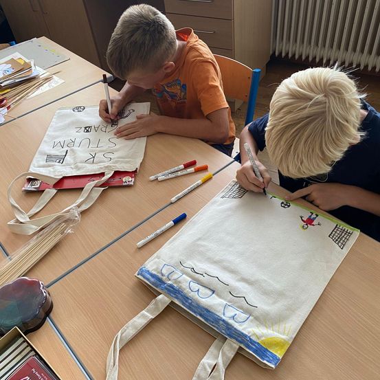 Zwei Jungen malen auf Leinwandtaschen in einem Klassenzimmer. Der Junge auf der linken Seite schreibt auf seine Tasche, während der Junge auf der rechten Seite ein Bild malt.