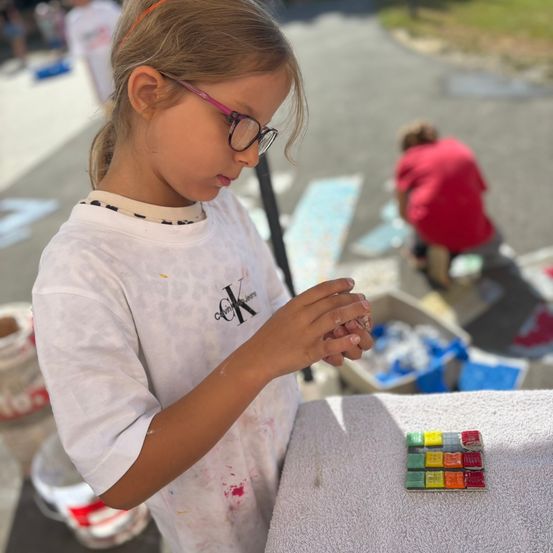 Ein junges Mädchen mit Brille und einem weißen Shirt mit CK-Logo schaut auf ein farbenfrohes Mosaikfliesen auf einem Tisch. Dahinter arbeitet eine Person in einem roten Shirt an einem blauen Design auf dem Boden.