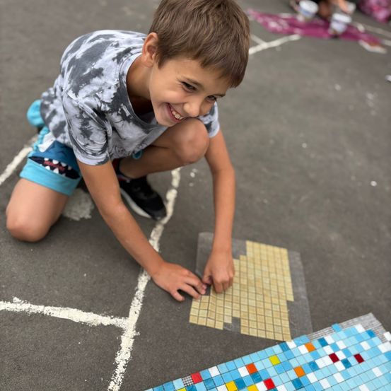 Ein junger Junge kniet lächelnd auf dem Boden und arbeitet an einem Mosaik mit bunten Fliesen. Er trägt ein Tie-Dye-Shirt und Shorts.