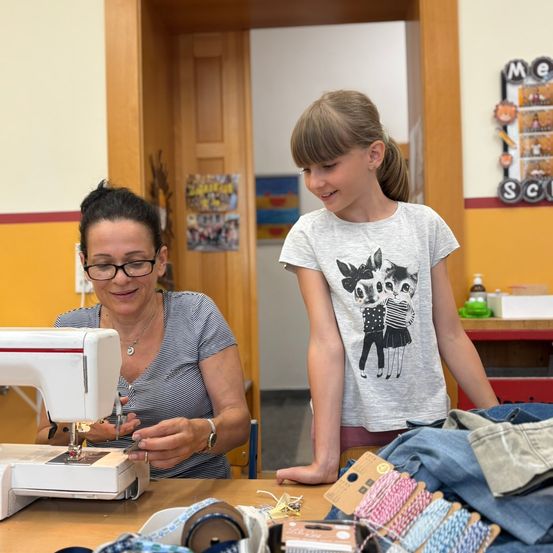 Eine Frau mit Brille und Armband näht an einer Nähmaschine. Ein Mädchen steht neben ihr und schaut sie an. Sie befinden sich in einem Raum mit einem mit Nähzubehör bedeckten Schreibtisch.