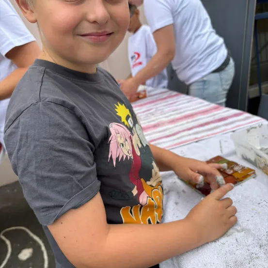 Ein junger Junge in einem grauen T-Shirt mit einem farbenfrohen Charakterdruck arbeitet an einem Bastelprojekt. Hinter ihm stehen Leute, einer hält einen Behälter. Der Tisch ist mit einem weißen Tuch mit rosa und weißen Streifen bedeckt.