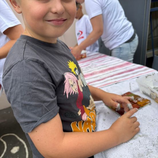 Ein junger Junge in einem grauen T-Shirt mit einem farbenfrohen Charakterdruck arbeitet an einem Bastelprojekt. Hinter ihm stehen Leute, einer hält einen Behälter. Der Tisch ist mit einem weißen Tuch mit rosa und weißen Streifen bedeckt.