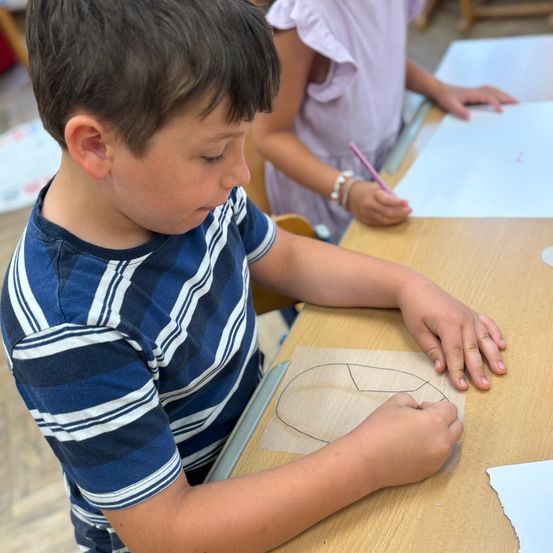 Ein Junge zeichnet auf einem Stück Holz, während er an einem Schreibtisch sitzt. Ein Mädchen sitzt neben ihm und schreibt auf Papier.