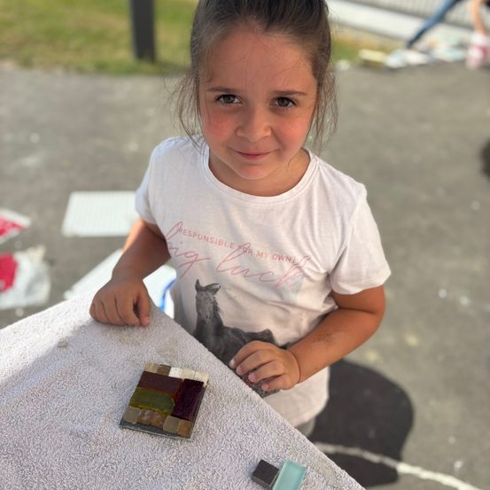 Ein junges Mädchen mit einem Lächeln arbeitet an einem Mosaikprojekt auf einem Tisch und trägt ein Shirt mit einem Pferd und Text.