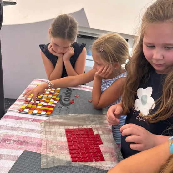 Drei junge Mädchen arbeiten an einem Bastelprojekt an einem Tisch und verwenden rote und mehrfarbige Fliesen. Eines der Mädchen hat die Hände im Gesicht, ein anderes konzentriert sich auf die Fliesen, und das dritte schaut zu.