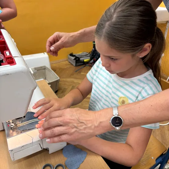 Ein junges Mädchen mit einem Pferdeschwanz und einem gestreiften T-Shirt benutzt eine Nähmaschine. Sie wird von einem Erwachsenen mit einer Uhr angeleitet. In der Nähe sind Scheren und Denim-Stücke auf einem Holztisch zu sehen.