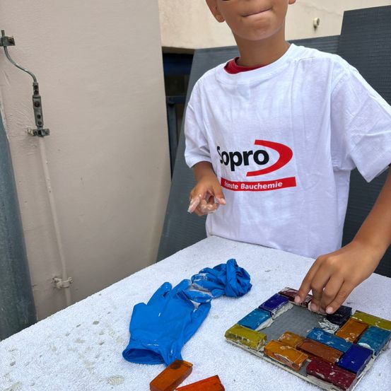 Ein junger Junge in einem weißen T-Shirt mit rotem Logo stellt ein Mosaik auf einem Tisch her. Er hat blaue Handschuhe und farbige Fliesen vor sich.