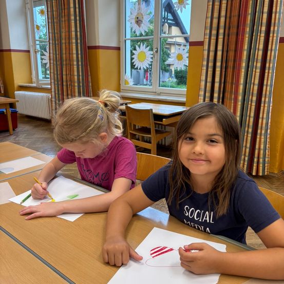 Zwei junge Mädchen sitzen an einem Tisch und sind mit dem Zeichnen beschäftigt. Eines der Mädchen zeichnet ein Herz, während das andere eine Blume zeichnet. Sie sind in einem Klassenzimmer mit Fenstern, die mit Sonnenblumen geschmückt sind.