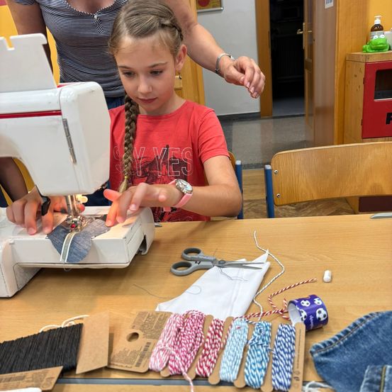 Ein junges Mädchen mit geflochtenen Haaren benutzt eine Nähmaschine, während eine Person hinter ihr ihre Hand hält. Verschiedene Nähmaterialien sind auf einem Holztisch verteilt.