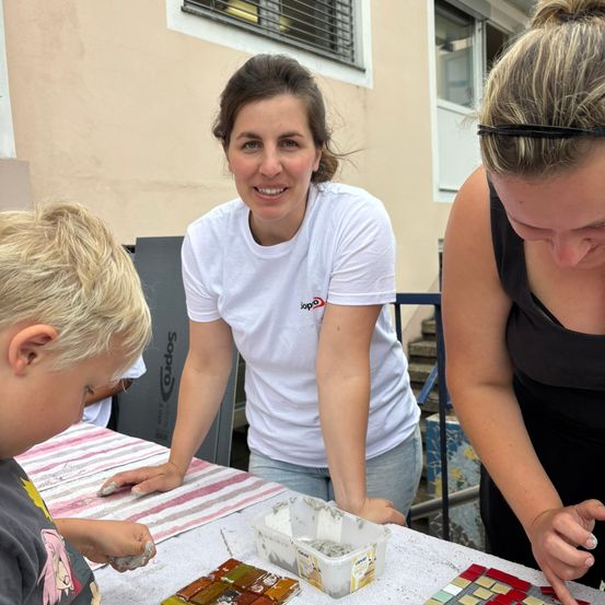 Drei Frauen und ein Junge arbeiten gemeinsam an einem Mosaikprojekt. Eine Frau trägt ein weißes T-Shirt, die andere ein schwarzes Top. Der Junge sitzt und trägt ein graues T-Shirt. Sie alle lächeln und konzentrieren sich auf ihre Aufgabe. Der Tisch vor ihnen ist mit einem weißen Tuch bedeckt, und es gibt bunte Fliesen auf dem Tisch.