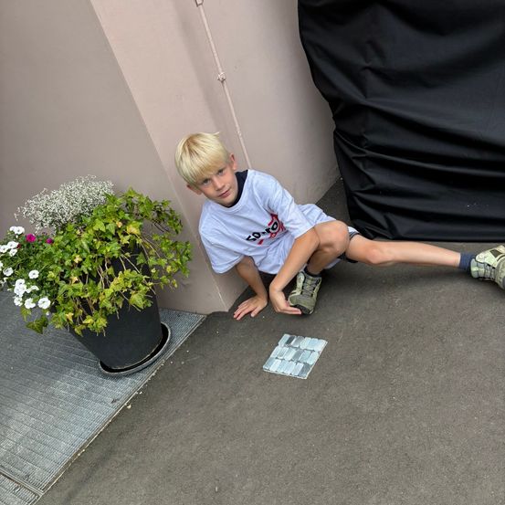 Ein Junge kniet neben einem kleinen Fliesenmosaik auf dem Bürgersteig, mit einem Topf neben ihm.