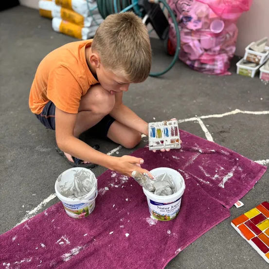 Ein junger Junge in einem orangefarbenen Shirt kniet auf dem Boden und hält eine Palette in der Hand, während er Farbe in zwei Eimern auf einem lila Handtuch mischt.