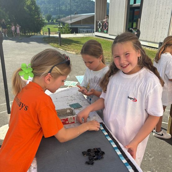 Drei Mädchen in weißen T-Shirts machen ein Bastelprojekt im Freien. Eine trägt ein orangefarbenes Shirt mit dem Wort 'Sonntage' darauf. Sie legen kleine Glasstücke auf eine Matte.