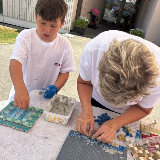 Ein Kind und ein Erwachsener arbeiten draußen an einem Mosaikprojekt, mit Fliesen und Mörtel auf einem Tisch.